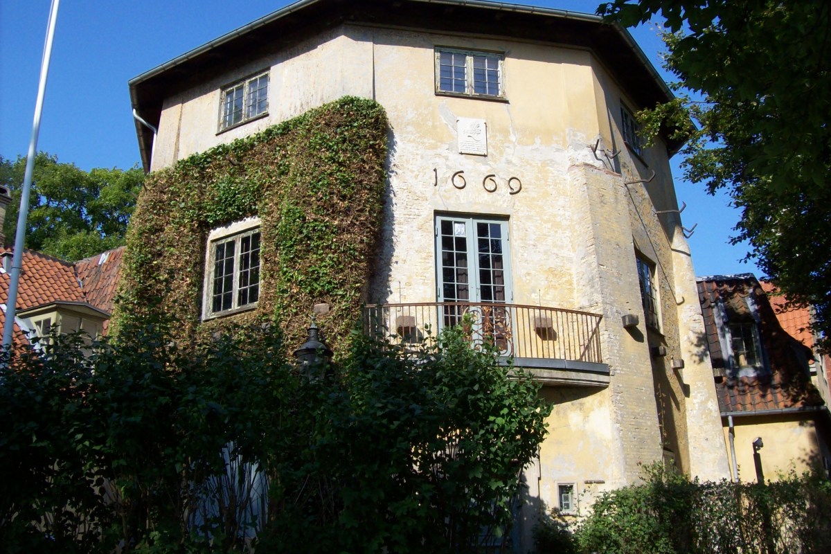 Exterior of Lille Mølle, a yellow ivy-covered tower house with balcony, dated 1669, among greenery.