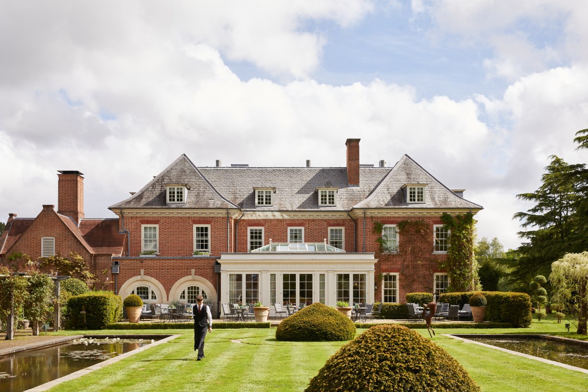 Red-brick manor hotel facade with gardens, hedges, pond and a staff member walking across the lawn