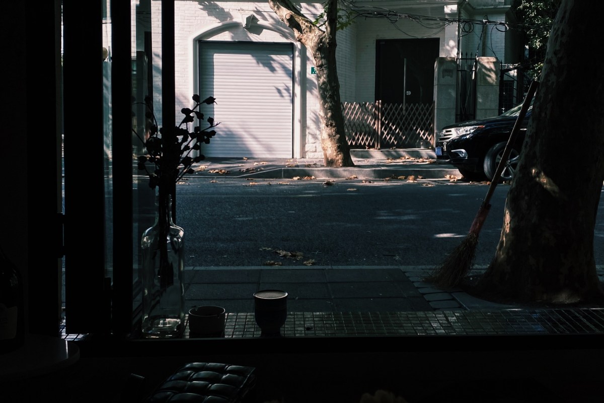View from inside a dimly lit restaurant looking out through glass doors to a quiet street with a vase of flowers on the sill