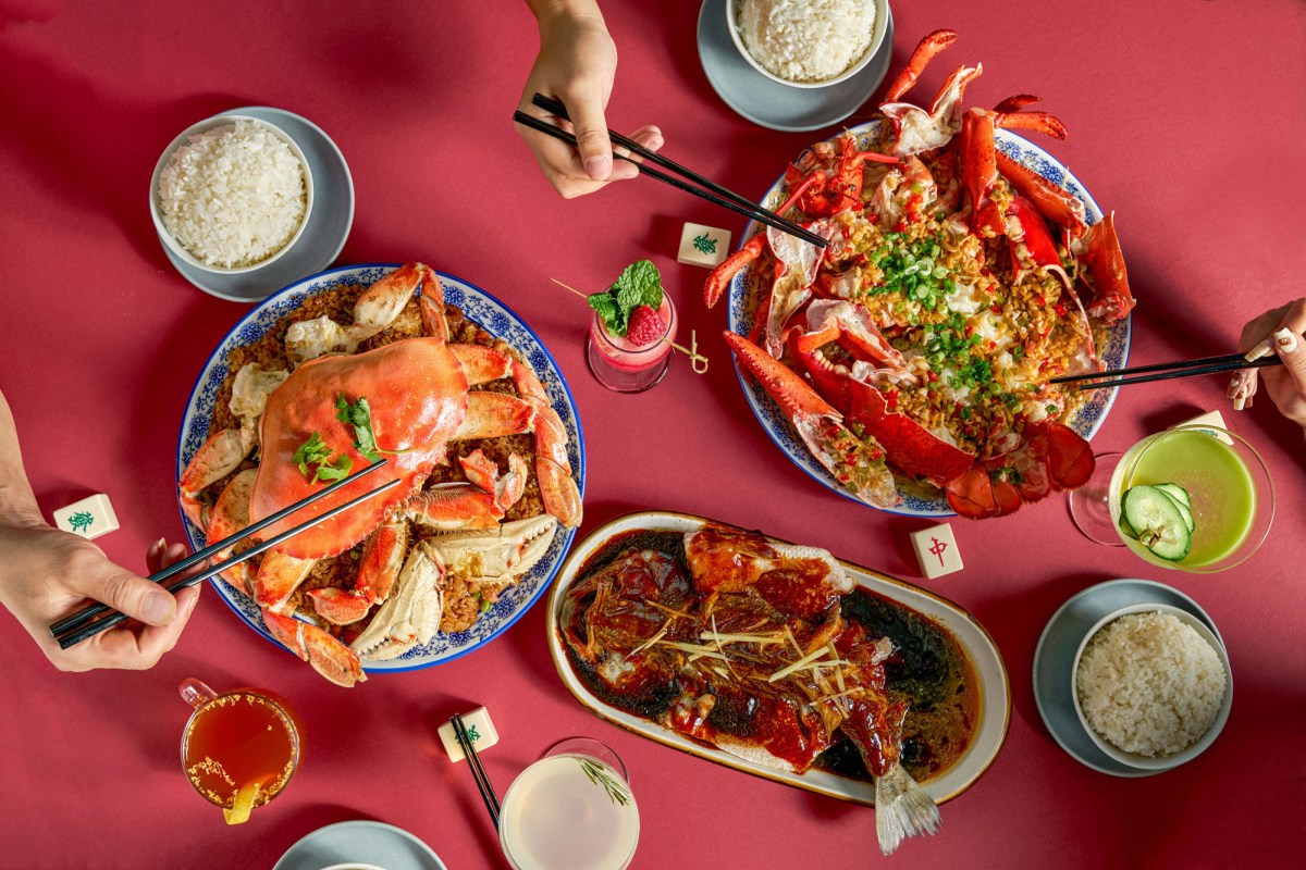 Overhead shot of a red table set with whole crab, lobster, fish, rice bowls and cocktails, hands reaching in with chopsticks