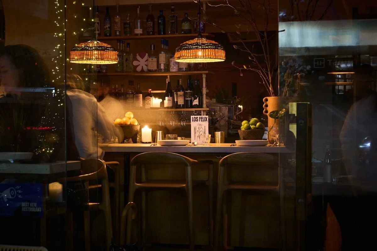 Exterior view through glass of Los Felix bar with illuminated pendant lights, bottles, and bar seating at night