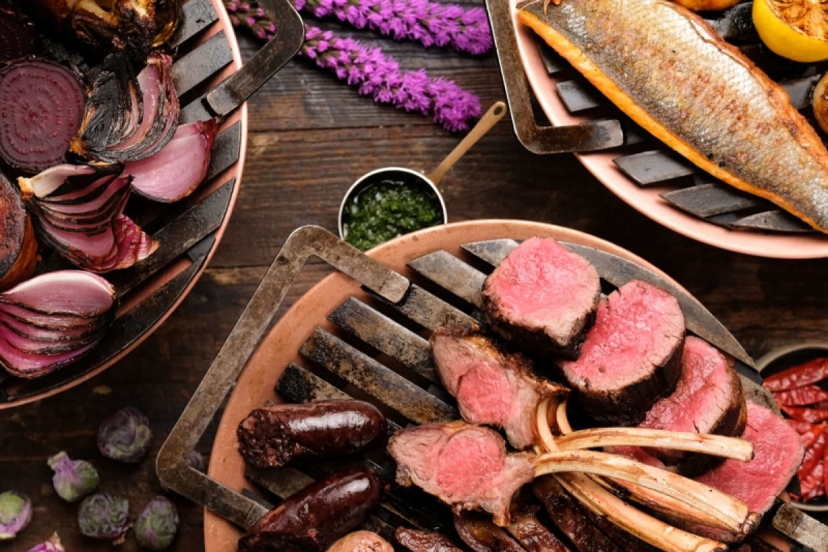 Overhead view of grilled meats, sausages, whole fish, and roasted vegetables on copper plates