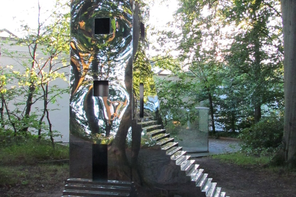 Polished stainless steel sculpture with geometric cutouts reflecting the surrounding woodland