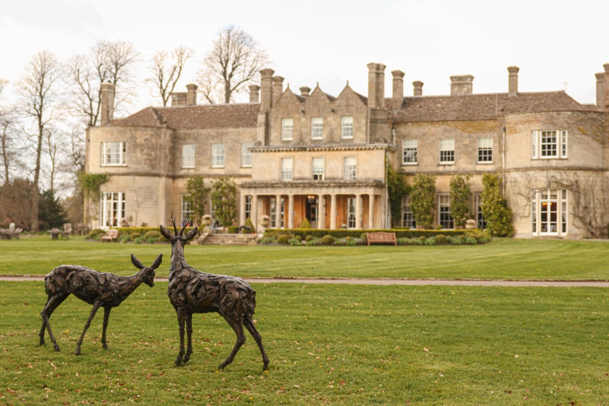 Grand stone manor house facade with chimneys and columned entrance, lawn with two bronze deer sculptures in foreground