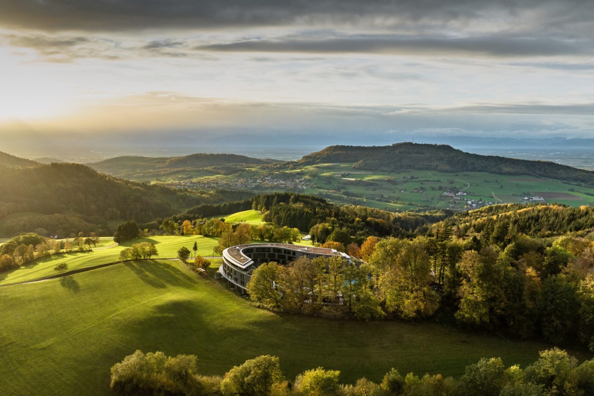 Aerial golden-hour view of the curved hotel building surrounded by forest and rolling green hills