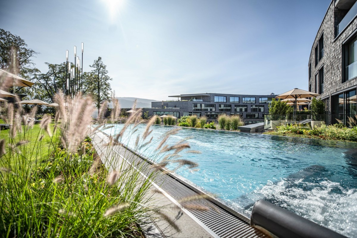 Infinity pool with hotel building and terrace loungers in background, surrounded by garden grasses
