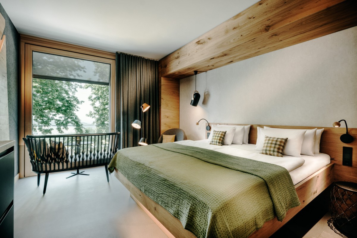 Modern hotel bedroom with wood paneling, green bedspread, and balcony view of trees