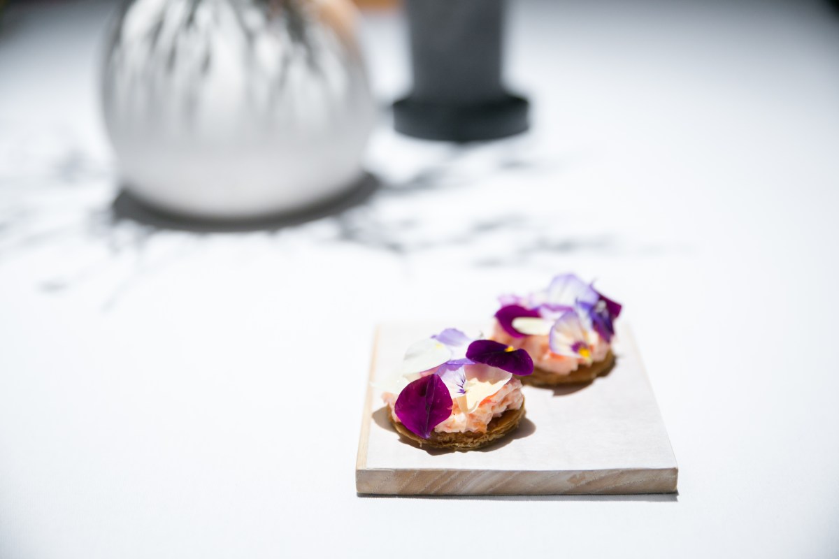 Two canapes topped with seafood and purple edible flowers on a wooden board