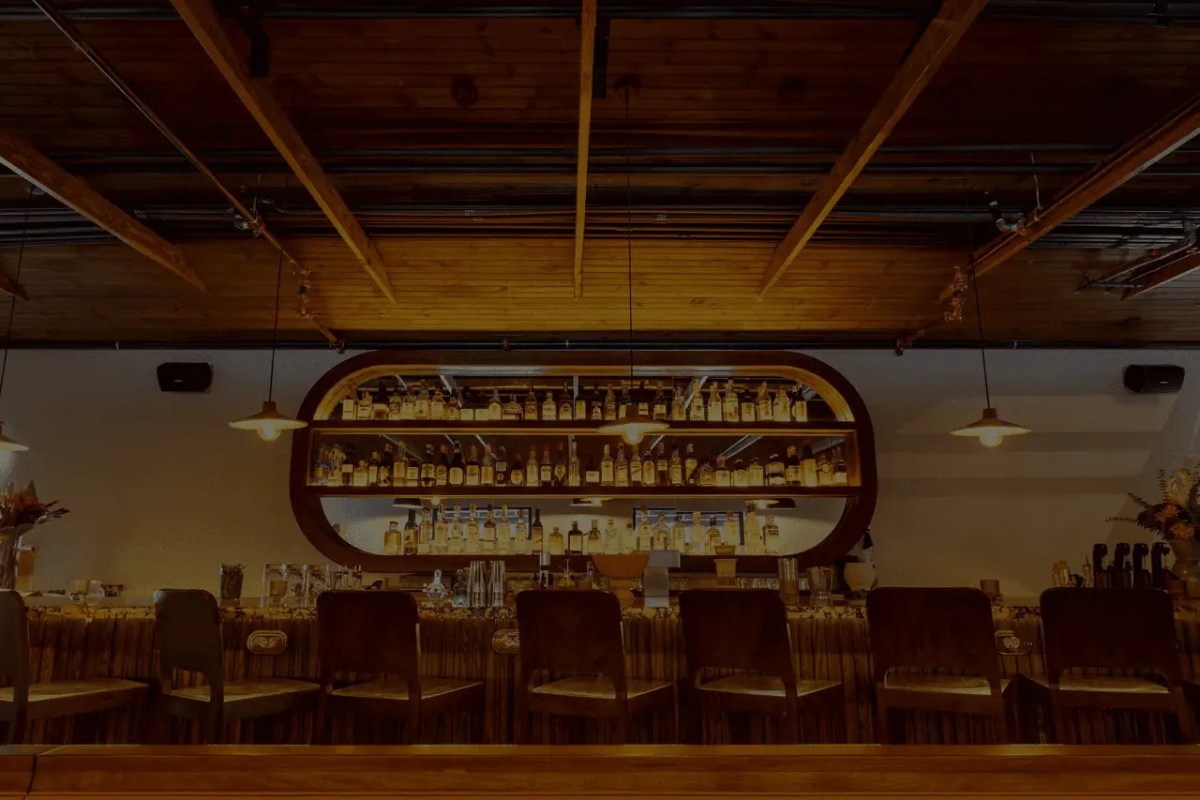 Wood-beamed bar area with shelves of liquor bottles and bar stools, warm lighting