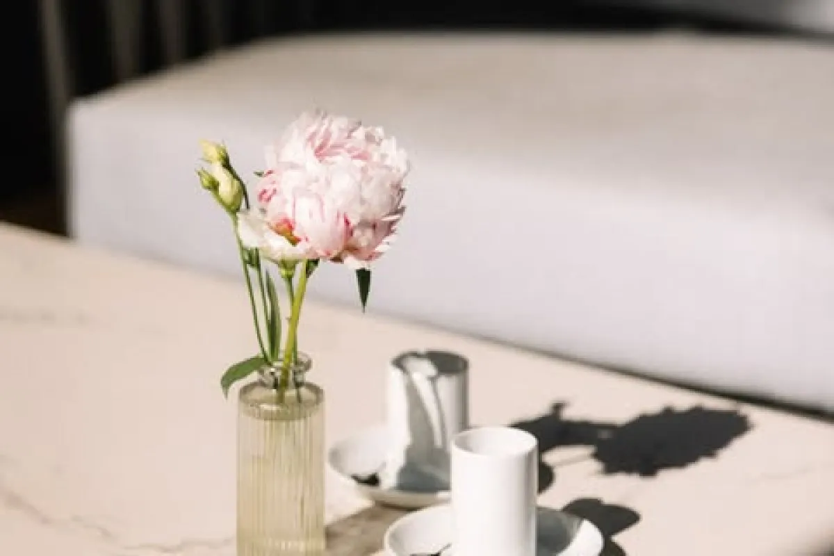 Close-up of a pink peony flower in a glass vase on a marble table with coffee cups