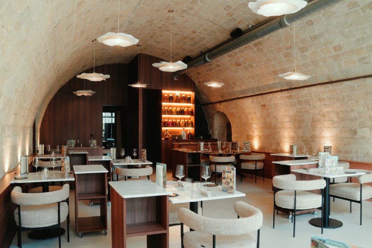 Vaulted stone dining room with wooden tables, white chairs, pendant lights and a backlit bar shelf