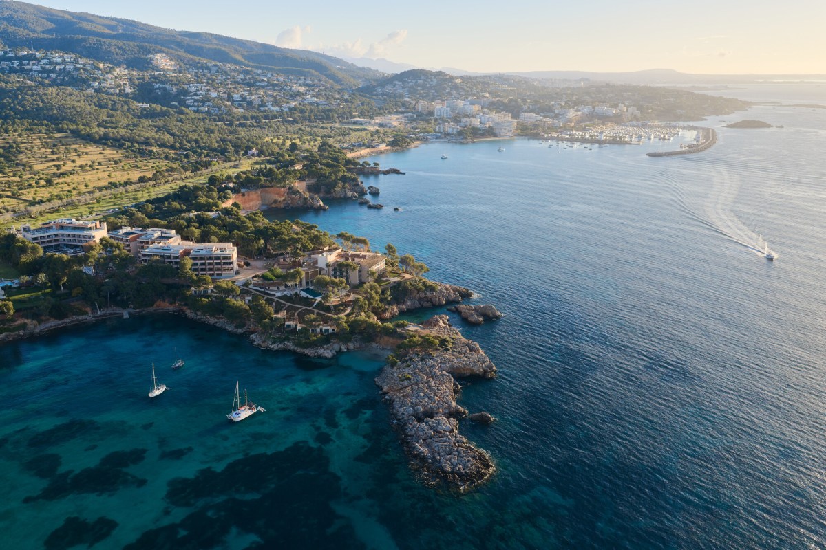 Aerial view of the hotel building nestled on a rocky coastline with turquoise sea, sailboats and surrounding hills