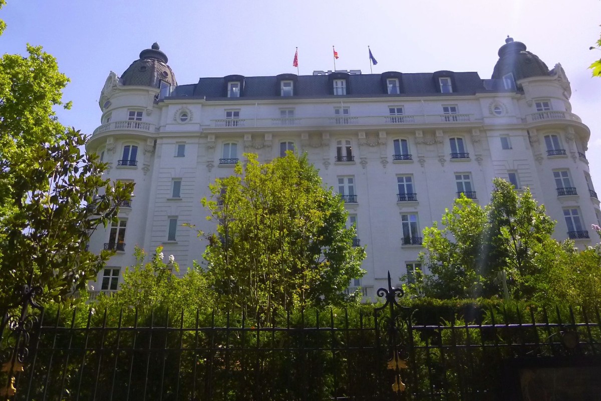 Daylight view of the white hotel building with twin domed towers, flags on roof, framed by green trees and a fence