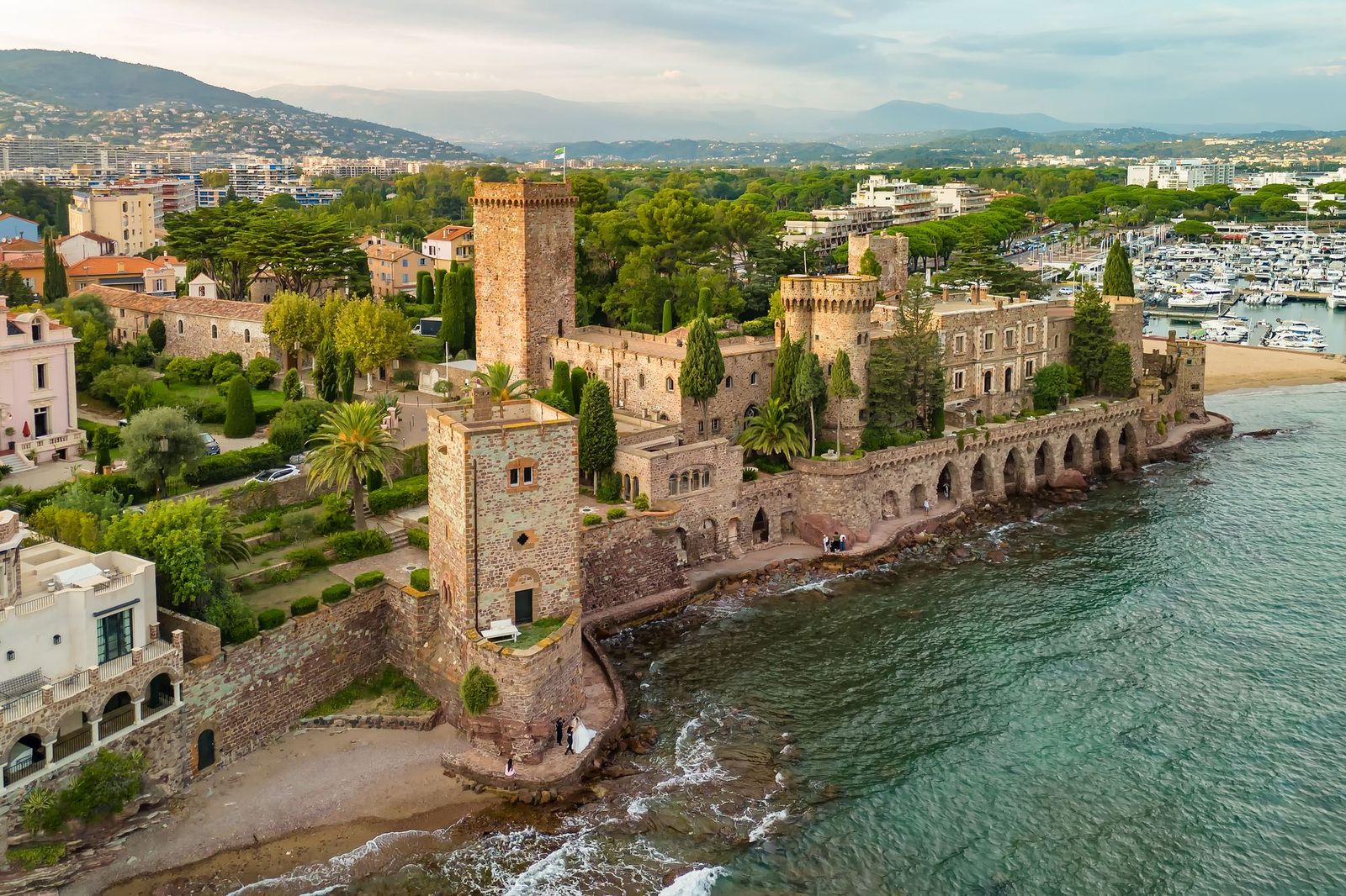 The Château de la Napoule from the air, its towers at the water's edge in Mandelieu-la-Napoule, with the marina and the hills beyond