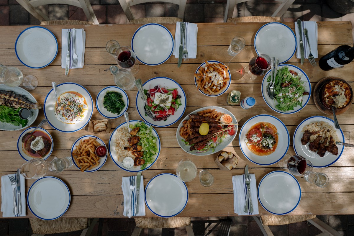 Overhead view of a wooden table laid with Greek dishes, salads, meats, and wine glasses