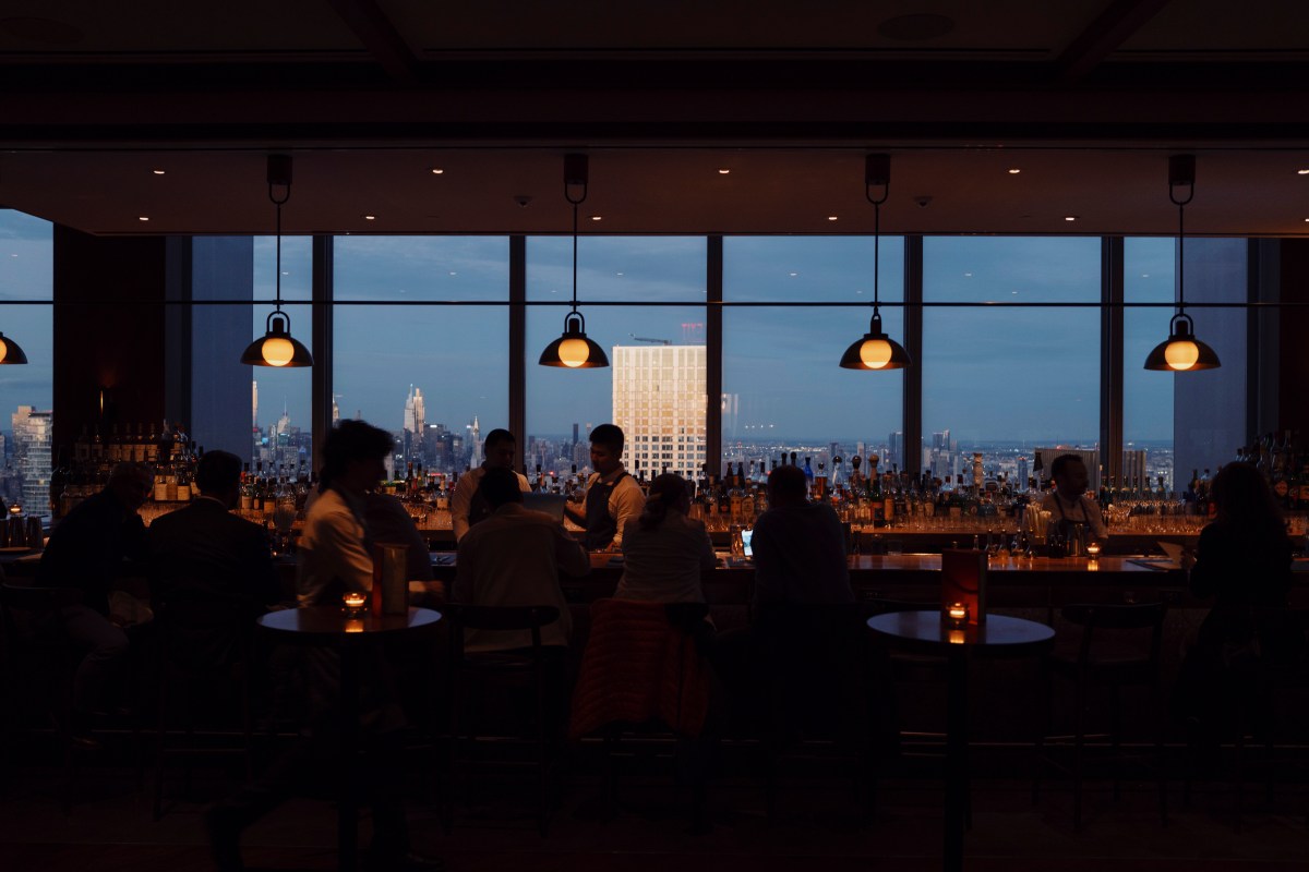 Dimly lit bar area with pendant lights, bottles, and a city skyline view through windows