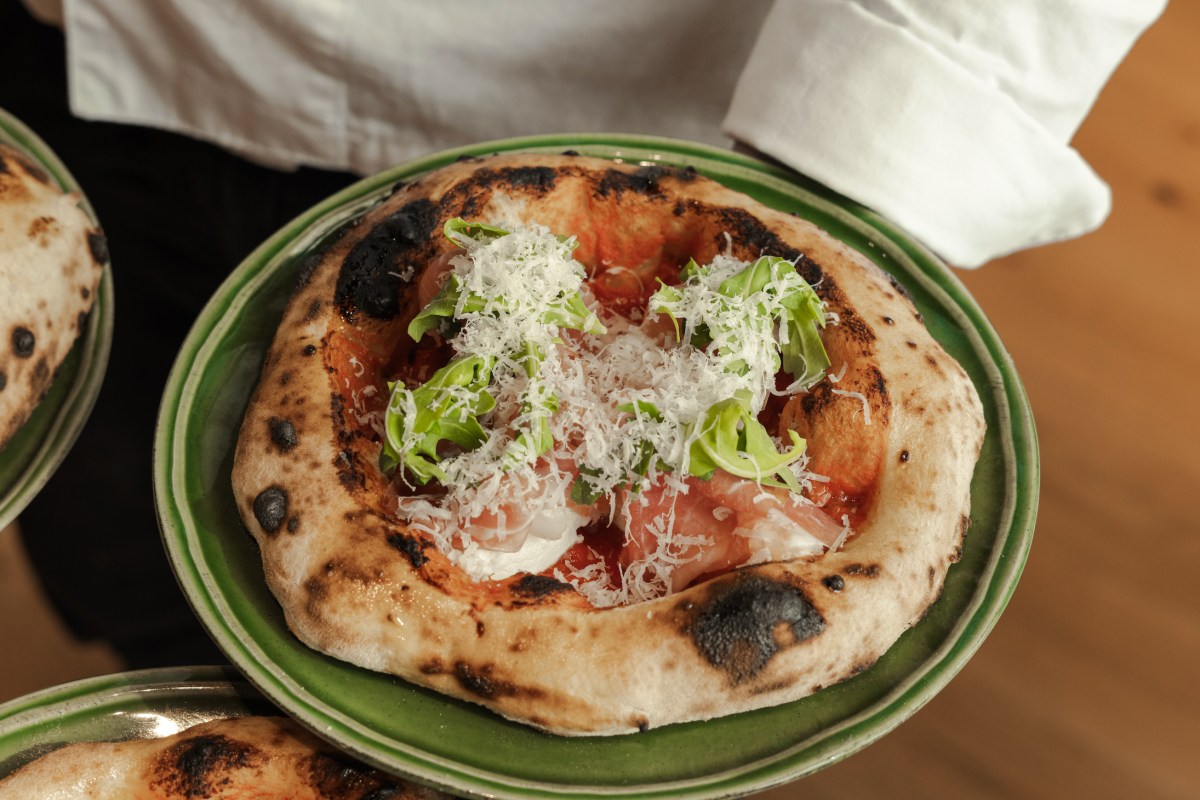 Overhead shot of a charred Neapolitan-style pizza topped with arugula, grated cheese, and prosciutto, held by a chef in white uniform