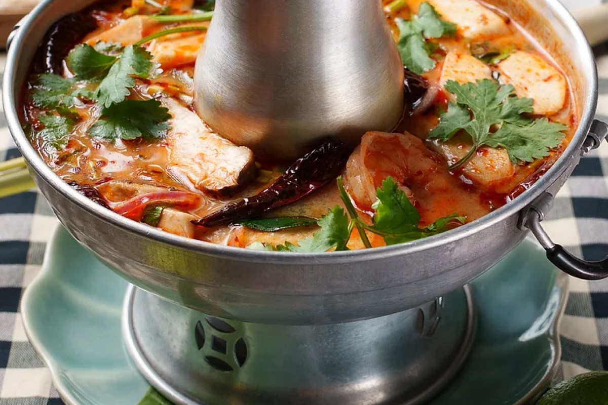 Close-up of tom yum soup with shrimp, fish, mushrooms, cilantro, and chili in a metal hot pot on a checkered tablecloth