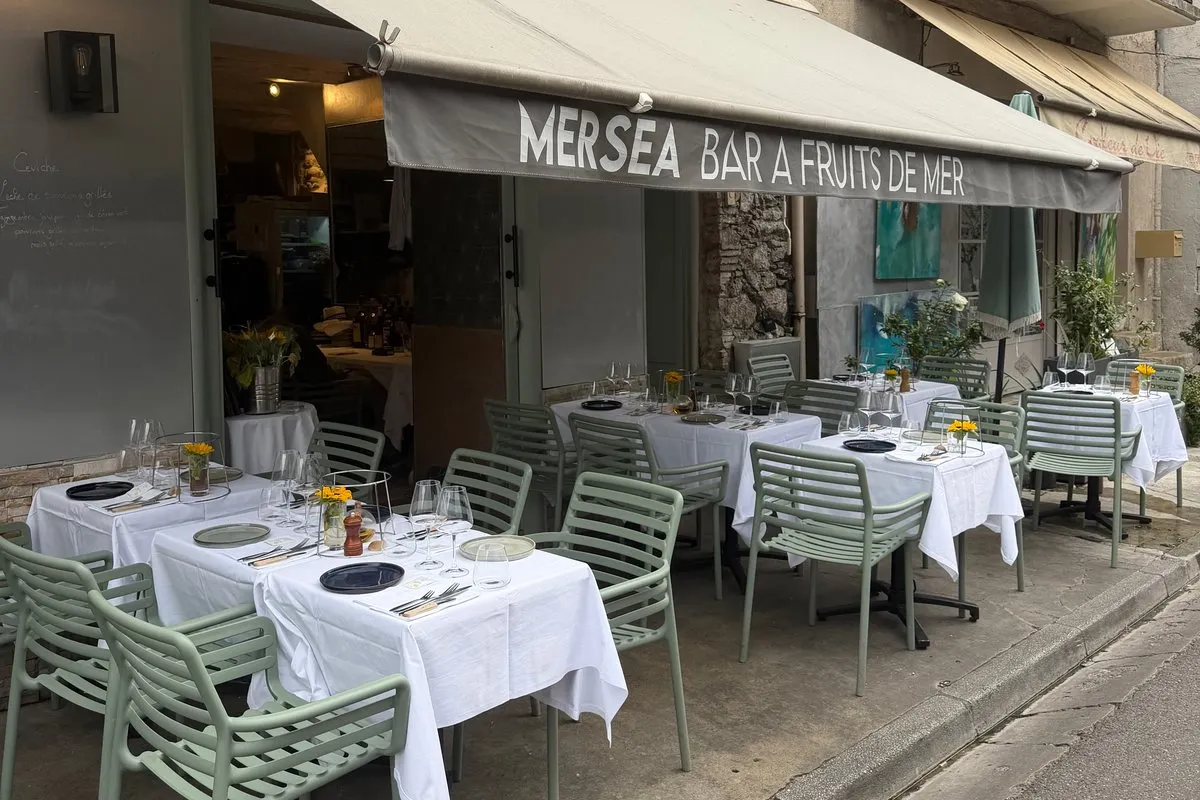 The terrace of Mersea on Cours Masséna in Vieil Antibes, with its grey awning reading «Mersea bar à fruits de mer», sage-green chairs and tables laid with white cloths and small yellow flowers