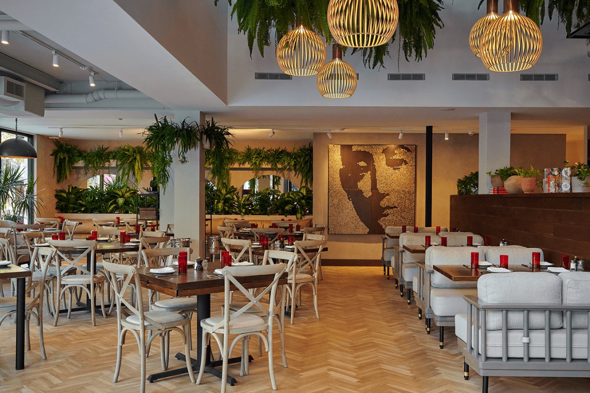 Interior dining room with wooden tables, cross-back chairs, hanging plants, pendant lights, and a mosaic face artwork on the wall
