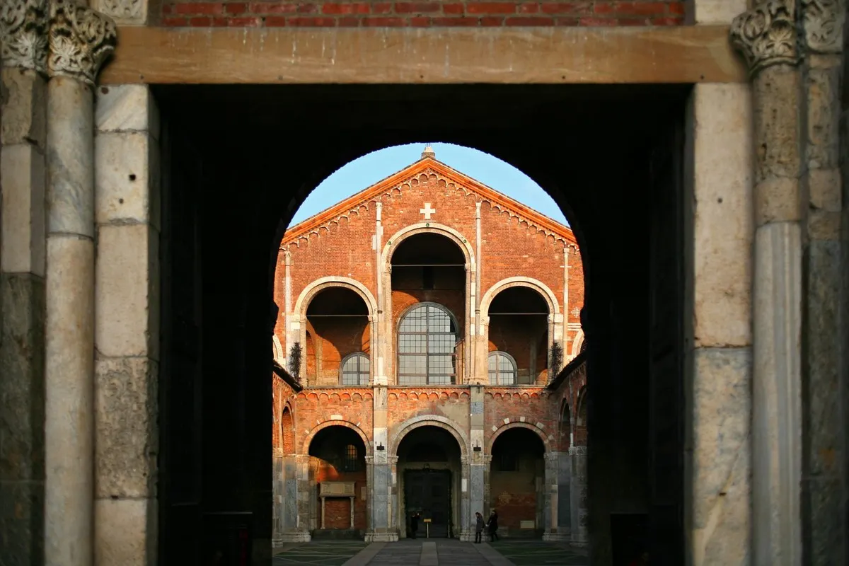 The brick Romanesque façade of the Basilica of Sant'Ambrogio in Milan, with its two tiers of arches, framed by the dark gateway of the atrium in warm evening light