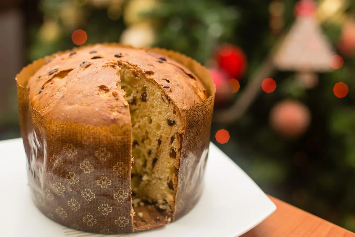 A tall Milanese panettone in its paper case, cut open to show the soft crumb with raisins, in front of a Christmas tree