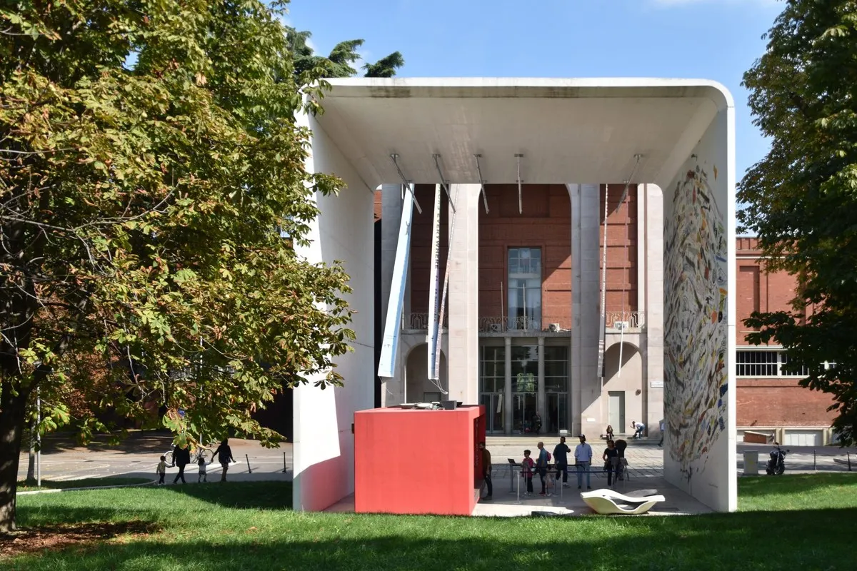 A white pavilion on the lawn of Parco Sempione in front of the brick Palazzo dell'Arte, home of the Triennale Milano