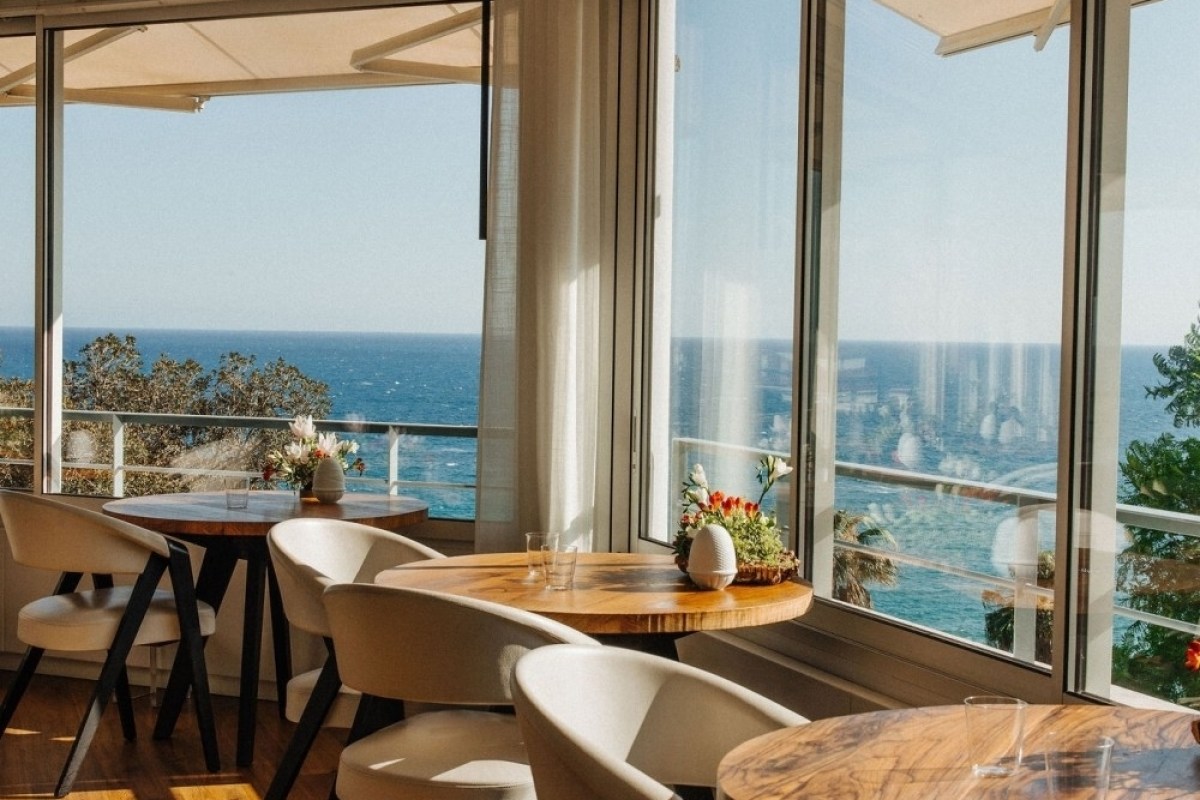 Restaurant dining room with round wooden tables, white chairs, and floor-to-ceiling windows overlooking the sea