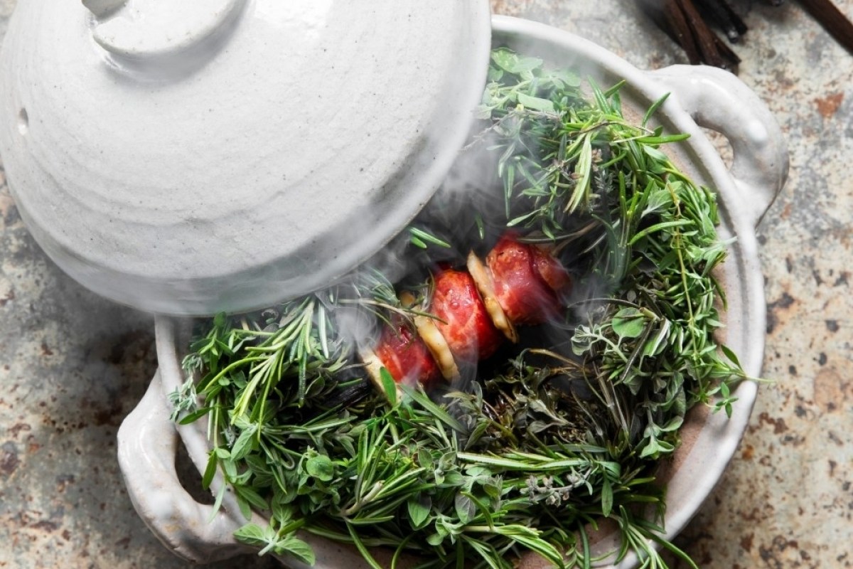 Steaming dish of wrapped meat and vegetables set in fresh herbs inside a ceramic pot with lid