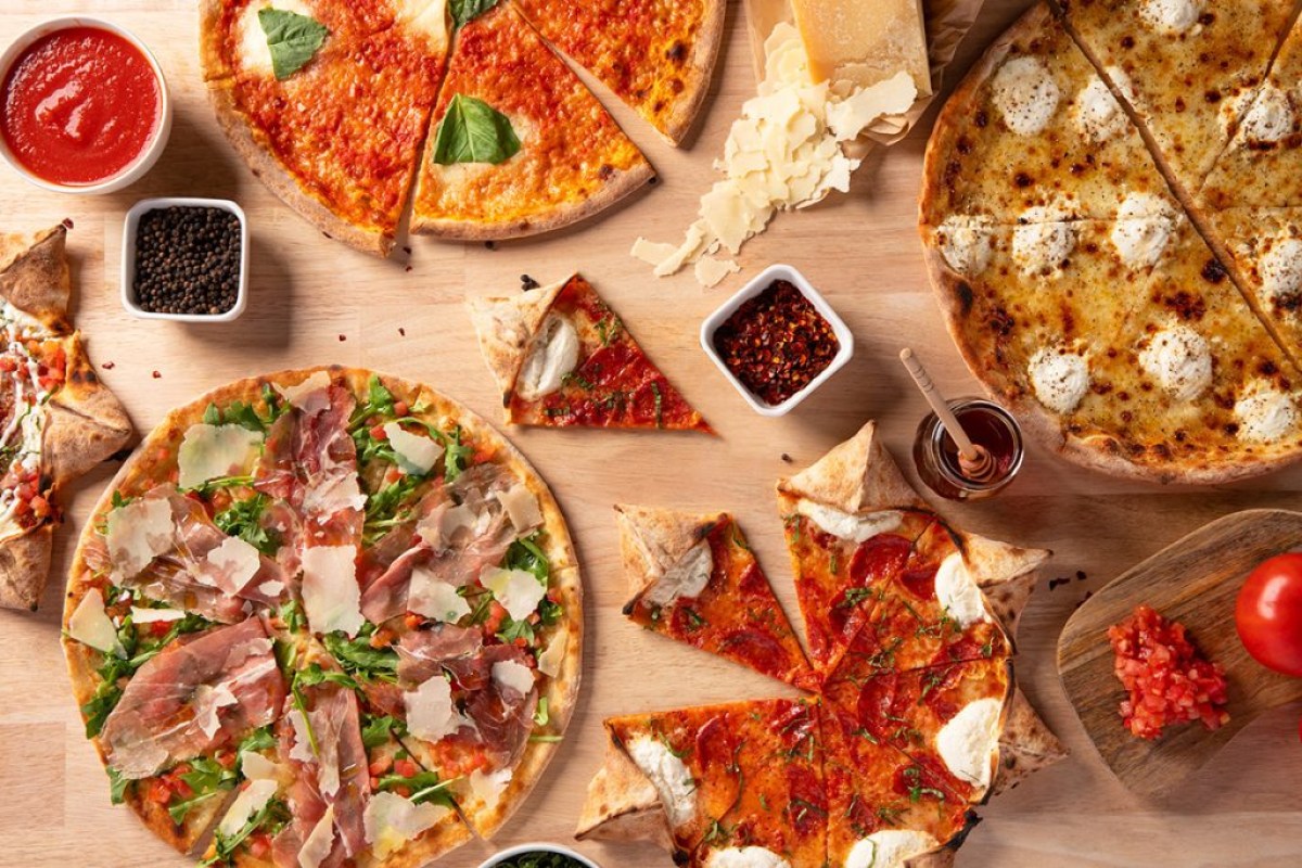 Overhead view of assorted sliced pizzas with toppings and condiments on wooden table