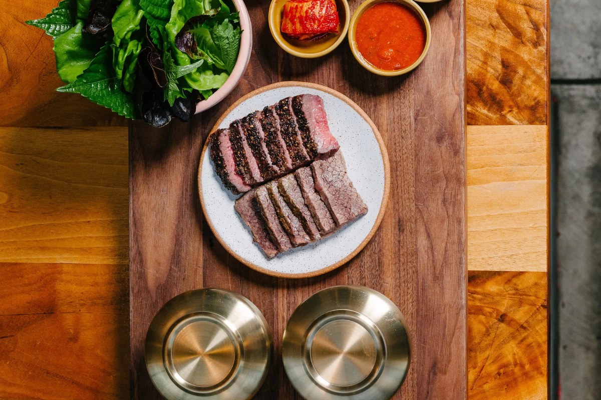 Overhead view of sliced steak on a plate with kimchi, sauce, and salad on a wooden board