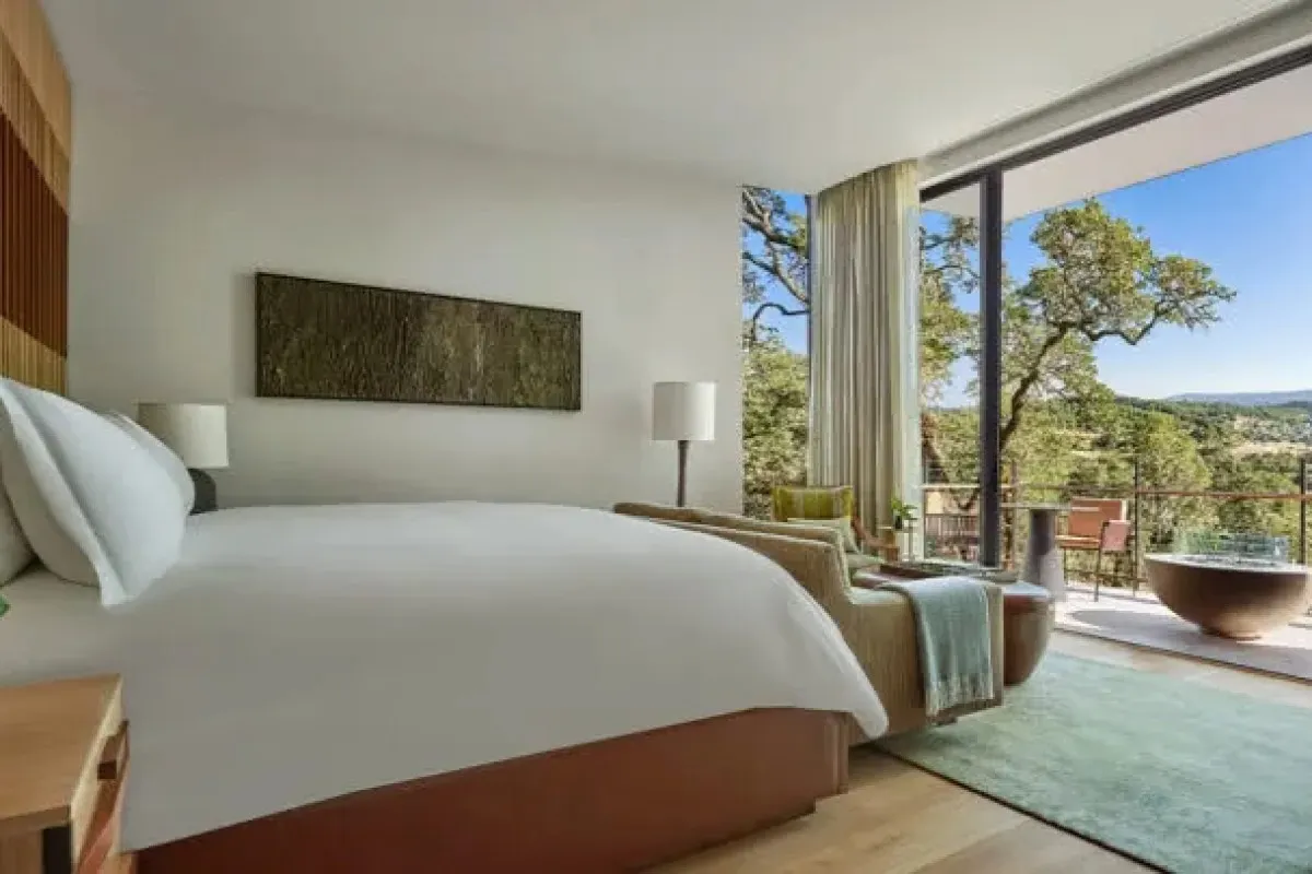 Bright guest room with white bed, armchair, large glass doors opening to a balcony with oak tree views
