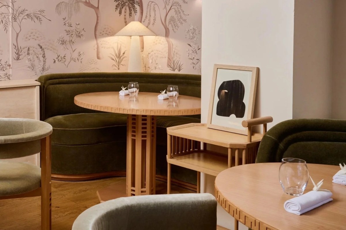 Restaurant dining room with round wooden tables, green velvet banquette, and patterned wallpaper