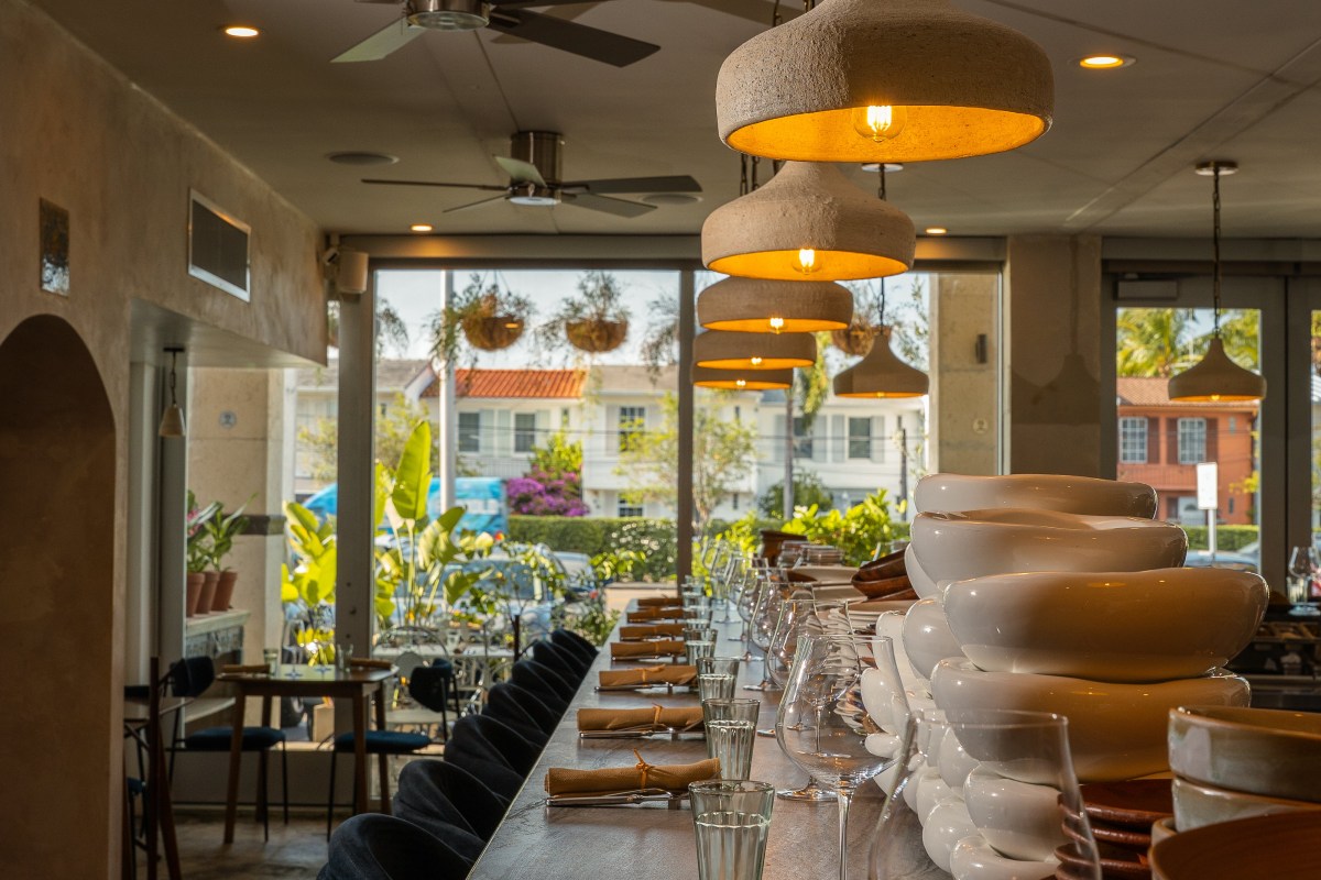 Restaurant interior with long bar, pendant lights, and view through windows to street and greenery