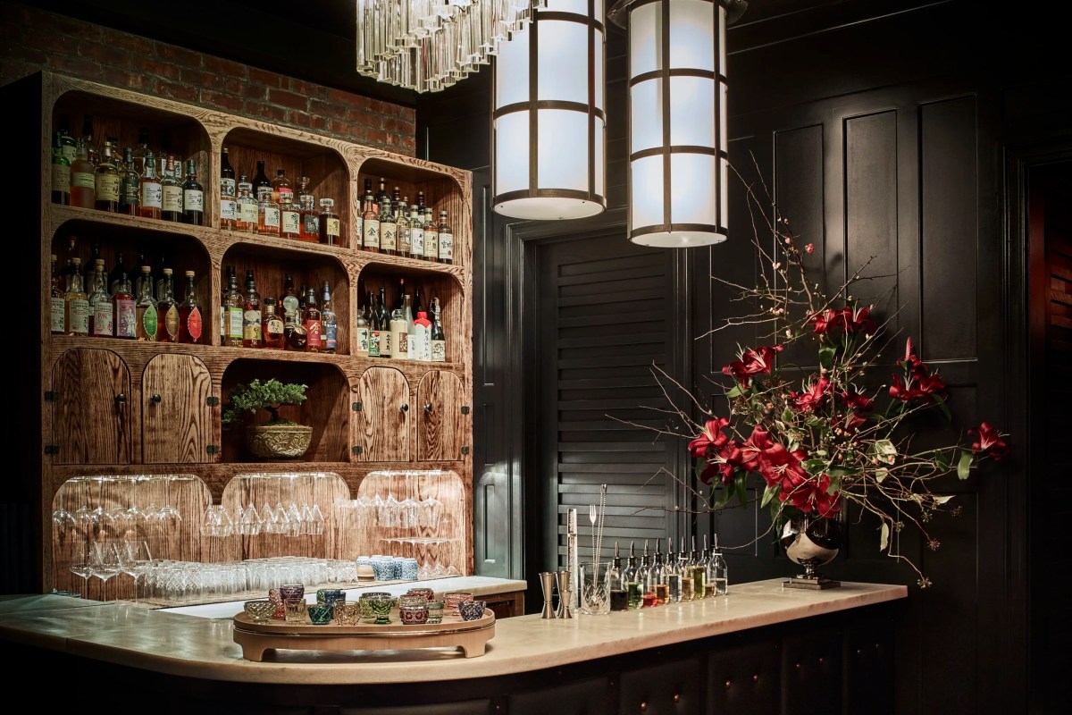 Close-up of restaurant bar with wooden shelving stocked with spirits, a bonsai, floral arrangement, and glass pendant lighting.