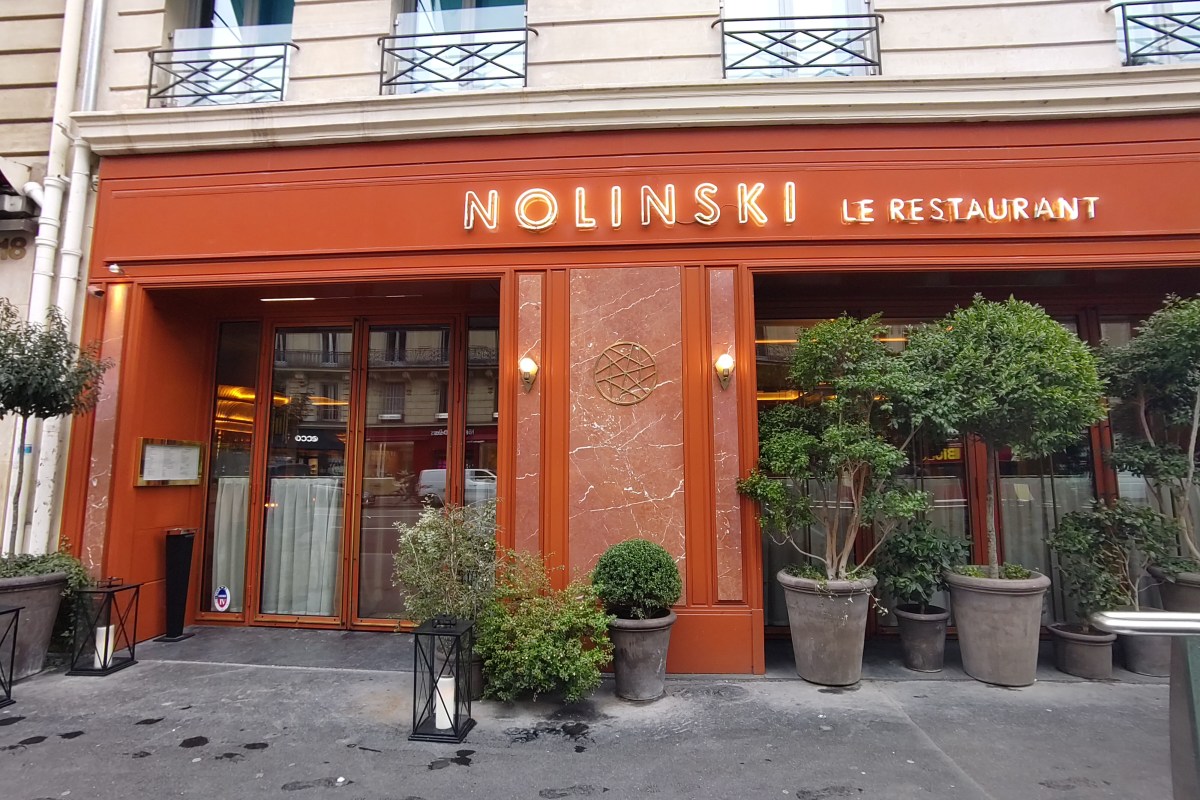 Exterior façade of Nolinski Paris hotel with orange signage, restaurant entrance and potted plants on sidewalk.
