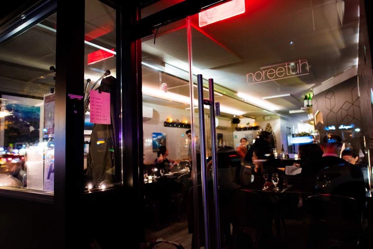 Exterior view through glass doors into Noreetuh's dining room at night, with neon sign and diners