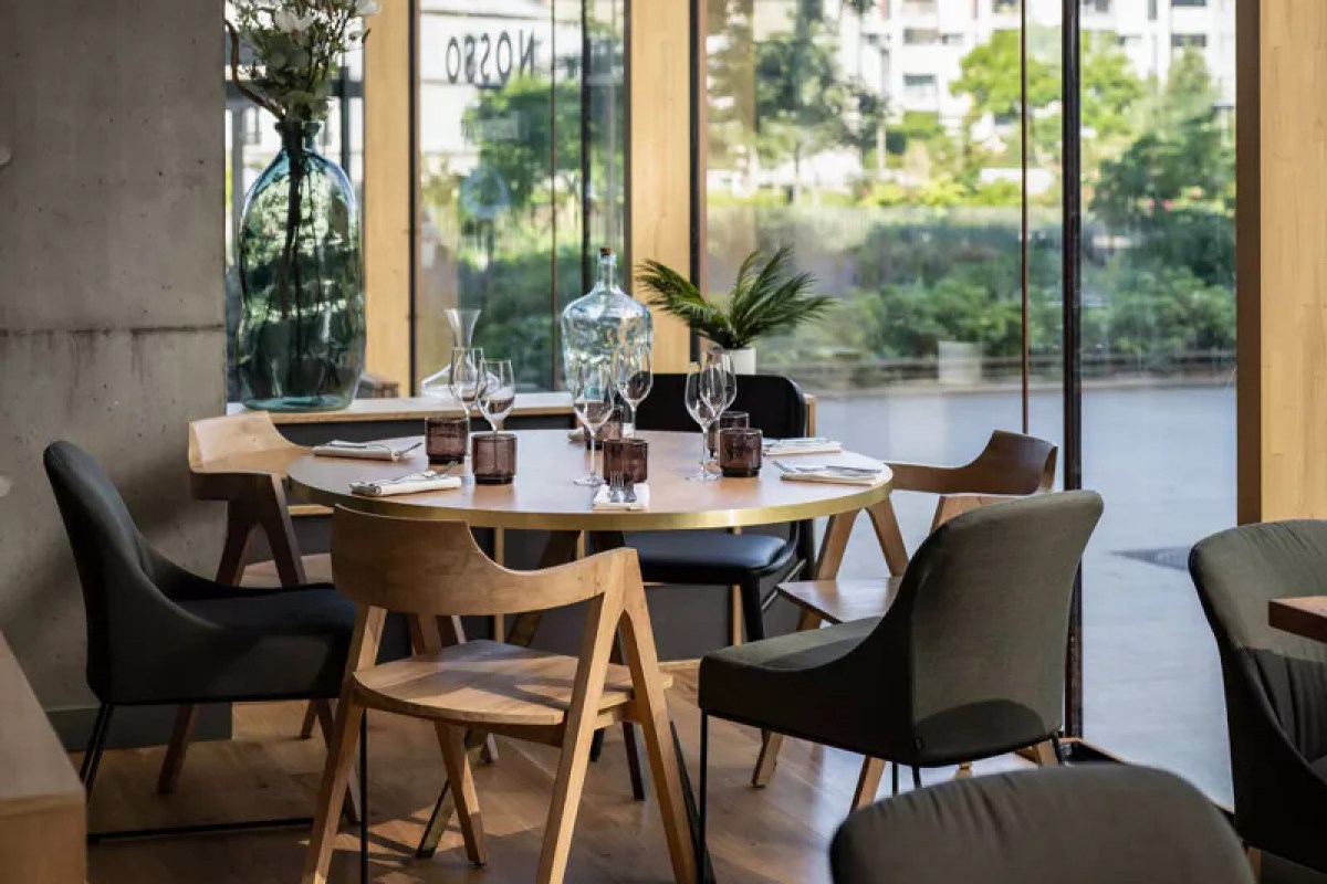 Round dining table set with glasses and carafes near large windows showing the NOSSO sign reversed, with greenery outside.
