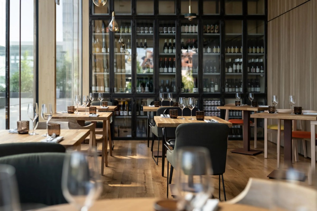 Restaurant dining room with wooden tables set for service, wine bottle display shelves, and large windows.