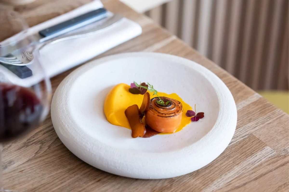 A plated dish with carrot puree, glazed carrot roll, and microgreens on a white ceramic plate at a restaurant table.