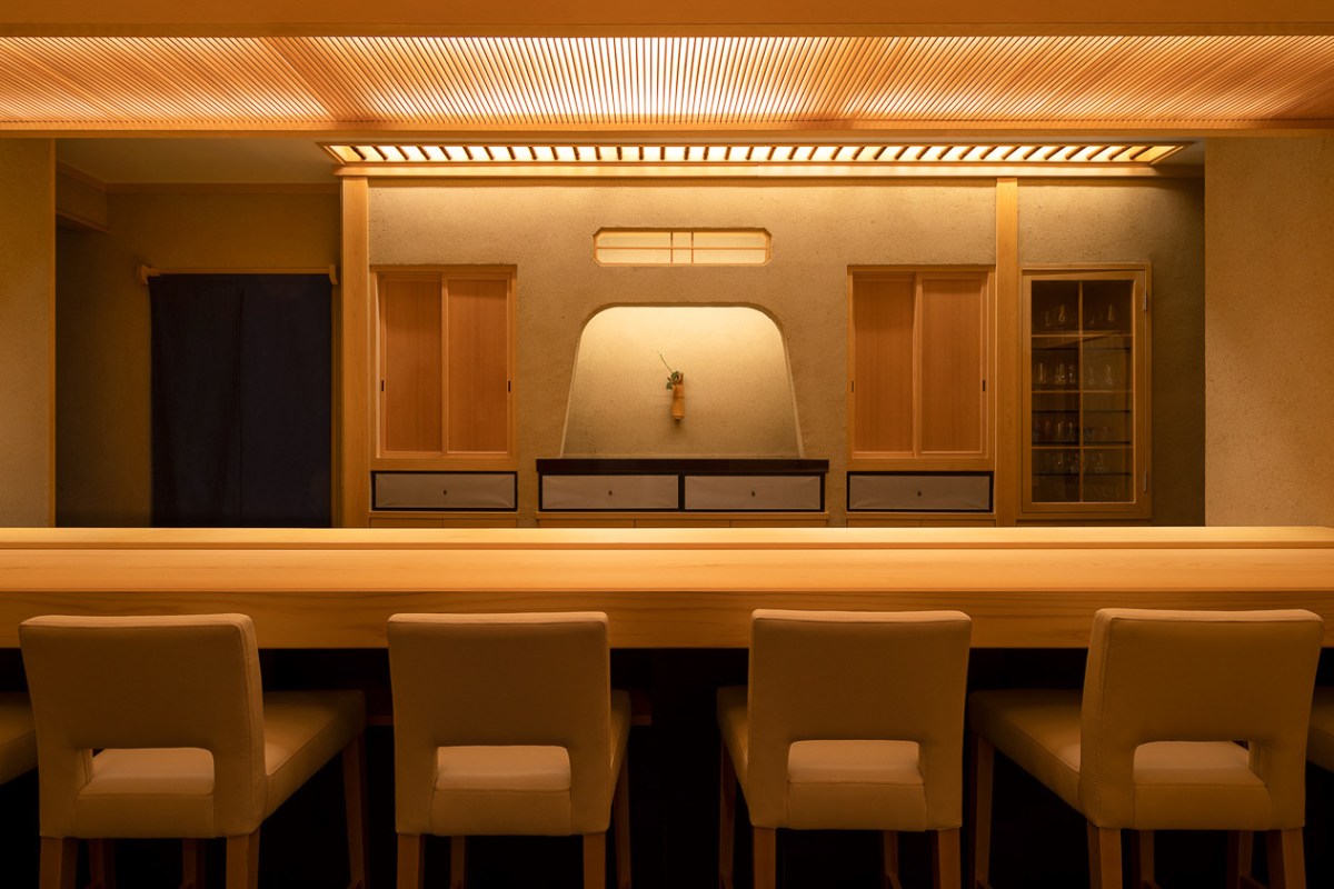 Empty wooden sushi bar counter with stools facing a minimalist Japanese-style back wall and cabinetry