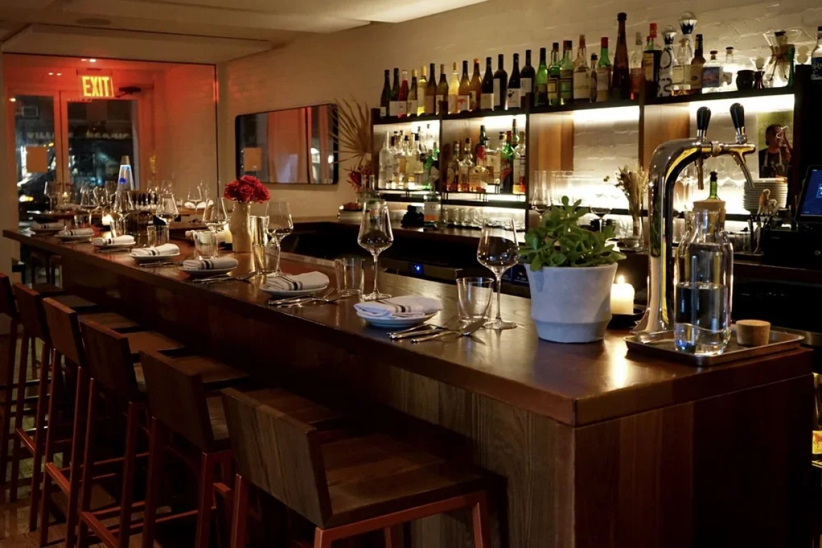 Dimly lit restaurant bar with stools, bottles on shelves, and set bar counter