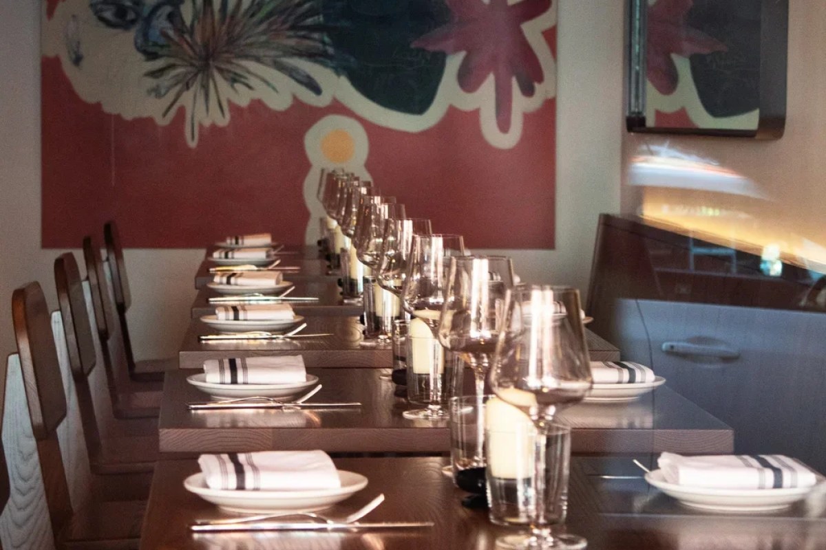 Long dining table set with wine glasses, napkins, and chairs, art on wall behind