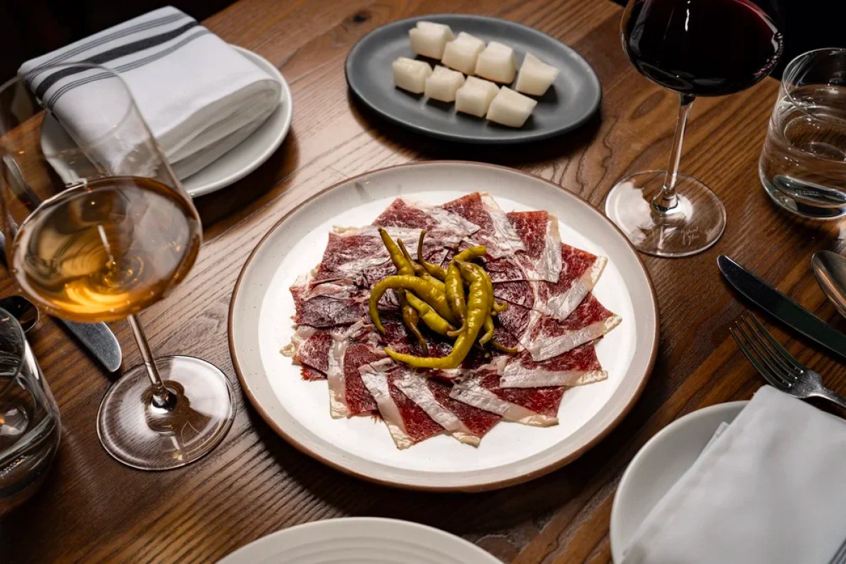 Plate of thinly sliced cured meat with peppers on a wood table with wine glasses