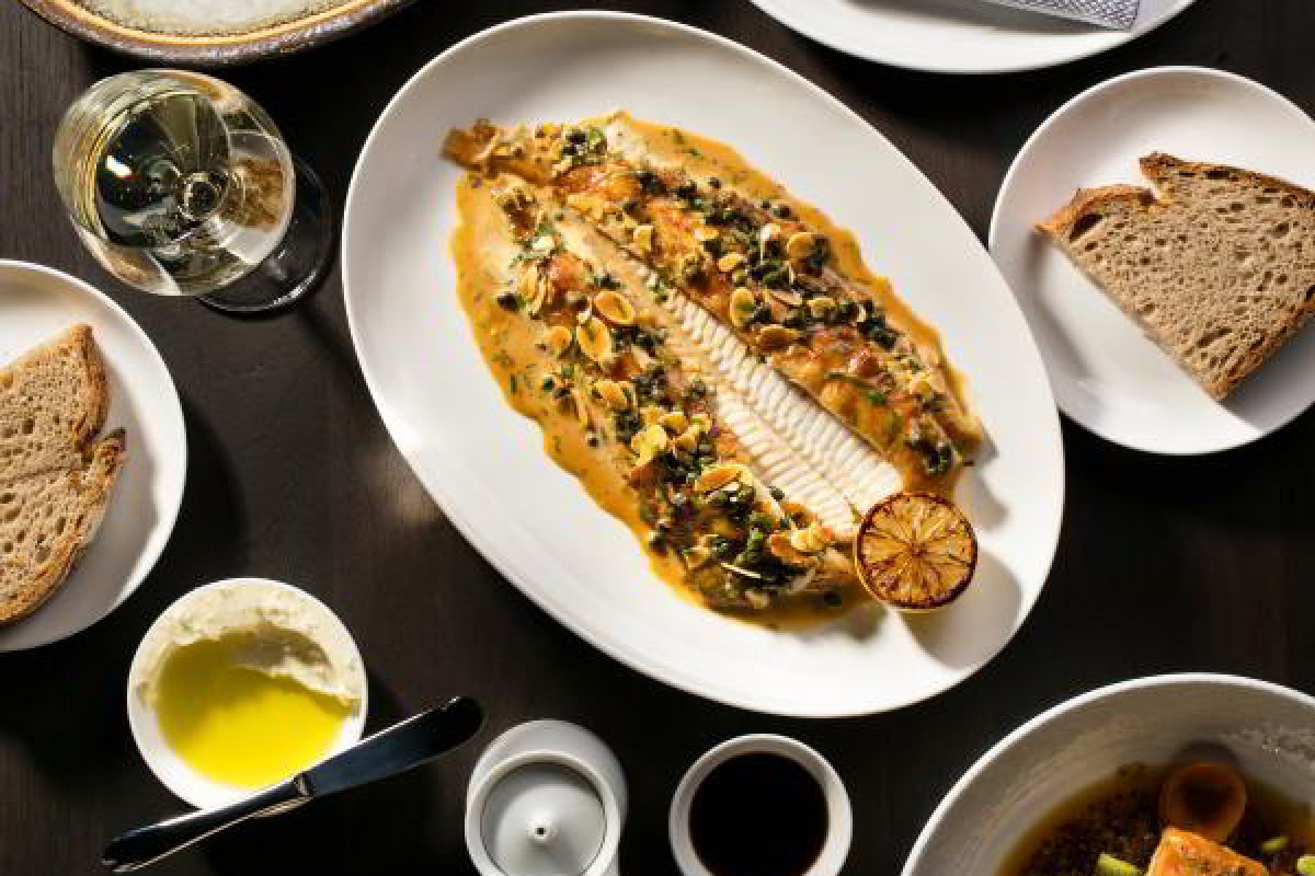 Overhead view of a whole roasted fish with caper sauce on an oval platter, surrounded by bread, oil, and condiment dishes