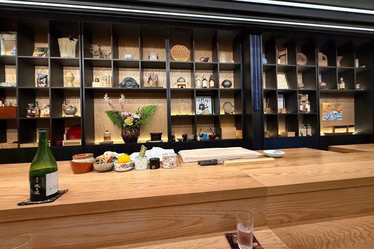 Sushi bar counter with sake bottle and condiments, backed by shelves displaying Japanese pottery and art