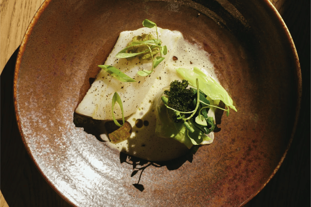 Overhead shot of a plated fish dish with greens on a textured brown ceramic plate