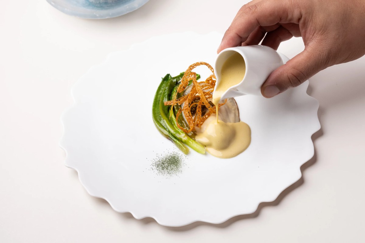 Overhead shot of a white plate with asparagus, crispy garnish, and sauce being poured from a small pitcher