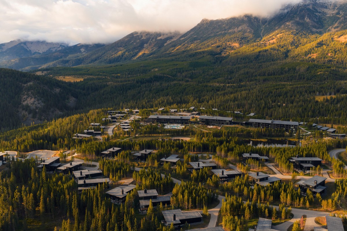 Aerial view of One&Only Moonlight Basin resort buildings and cabins nestled in forest below mountain peaks at sunset.