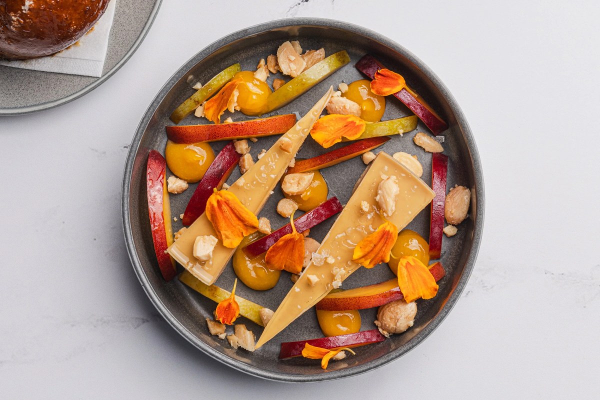 Artfully plated dessert with thin caramel shards, fruit slices, orange flower petals, and nuts on a dark round plate